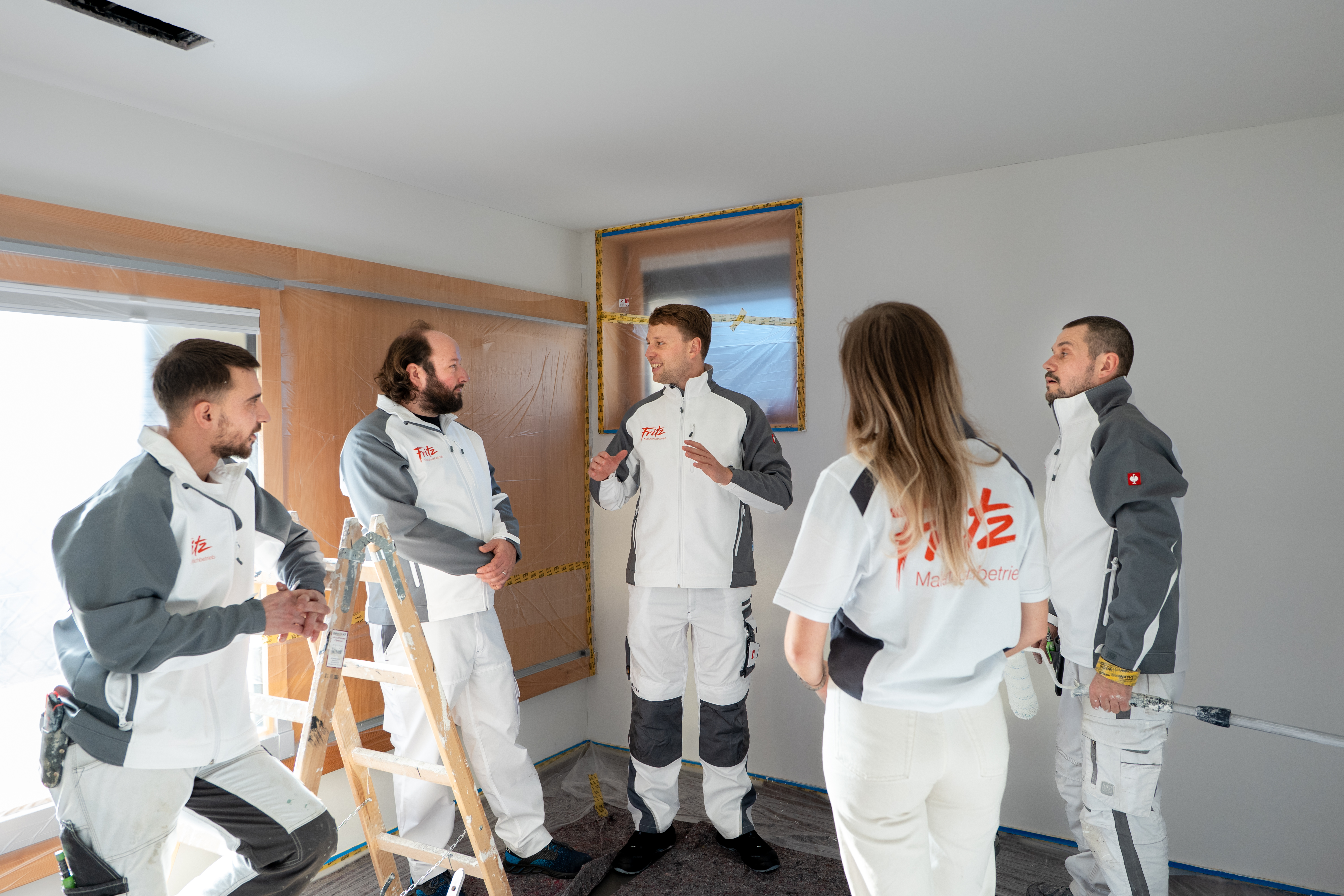 Fünf Mitarbeiter eines Malerbetriebs besprechen geplante Malerarbeiten in einem vorbereiteten Zimmer