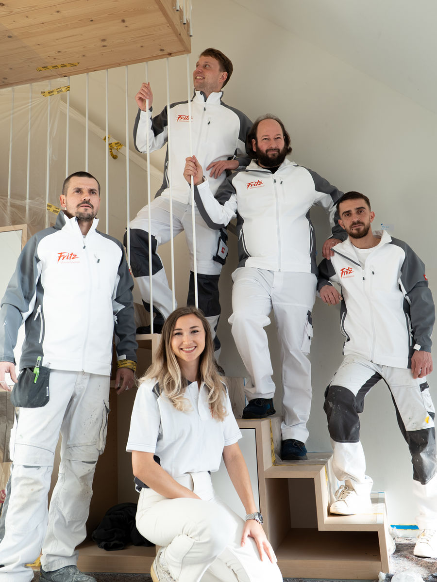 Six smiling craftspeople in white and grey workwear pose on a wooden staircase in a new building