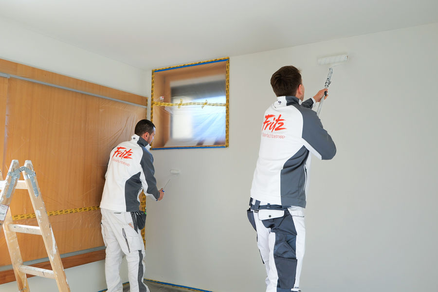 Two painters in Fritz painting company workwear paint the walls of a room, with windows and floor masked off